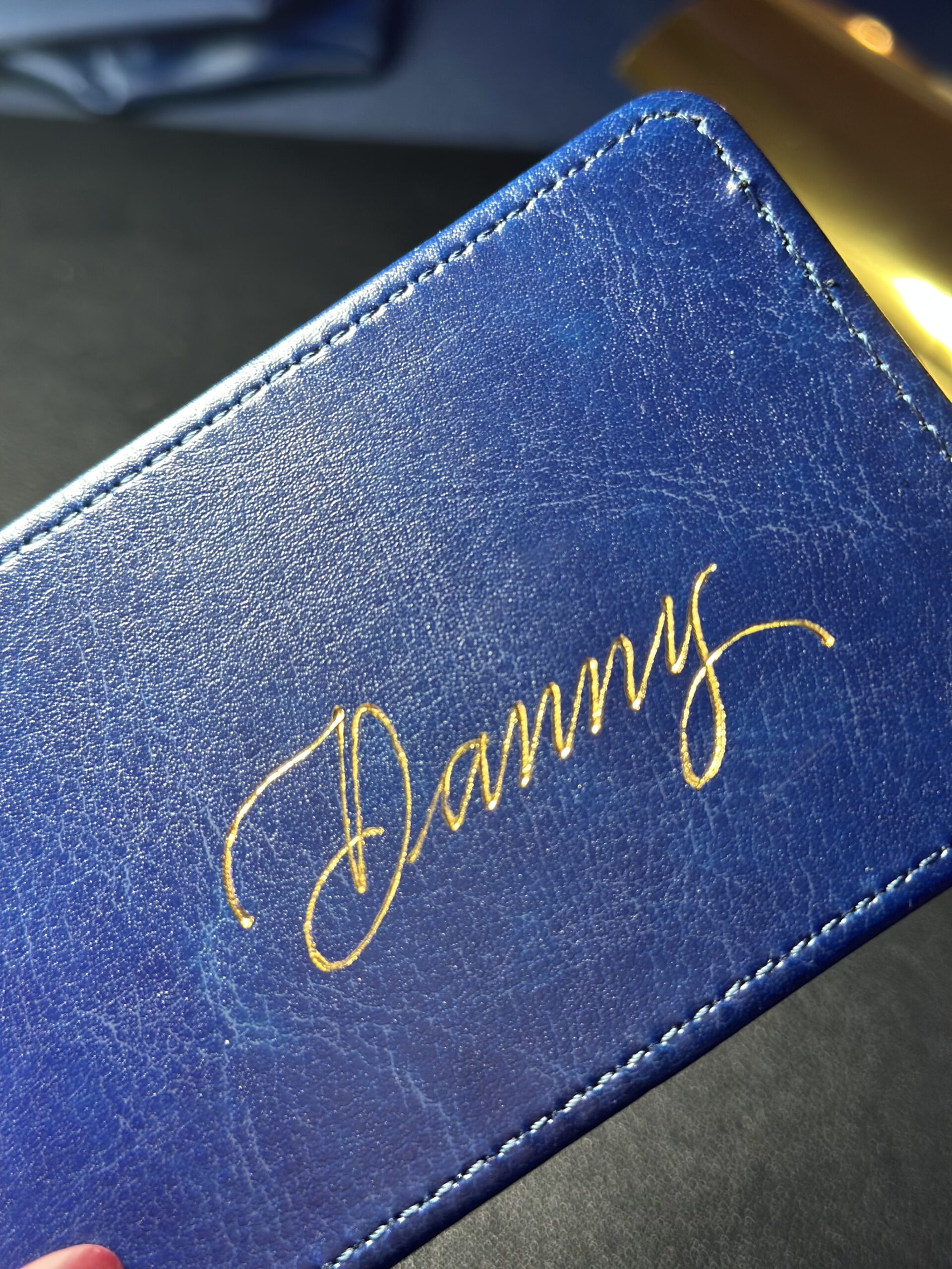 Blue leather luggage tag personalized with the name 'Danny' in shiny gold foil during a live event.